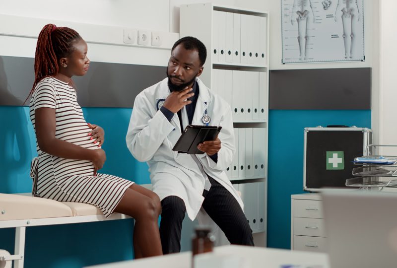 Male obstetrician taking notes on digital tablet at checkup visit, talking to patient with pregnancy belly in medical office. Doctor consulting expectant woman with baby bump to give advice.